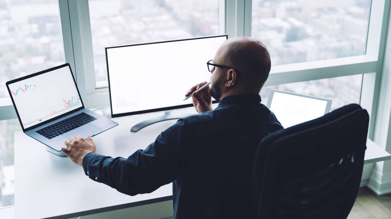 homem de costas para a foto com três telas de computador estudando atentamente