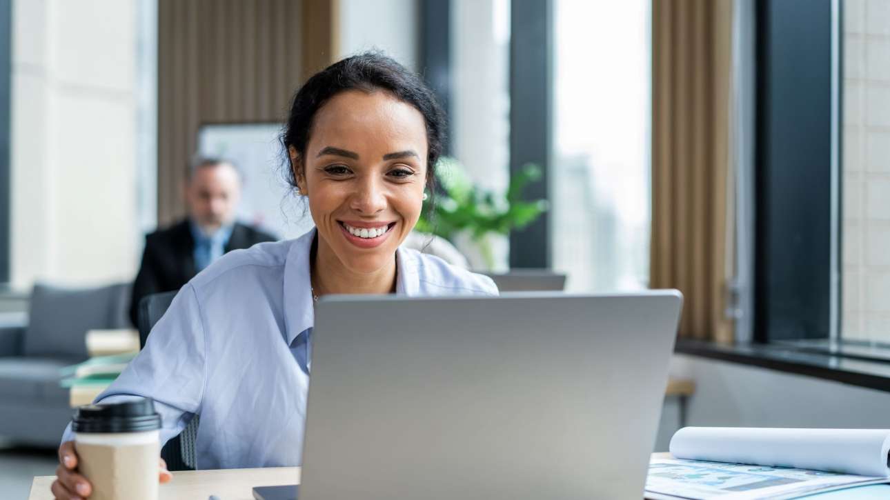 mulher em frente a um computador, sorringo e segurando um copo de café