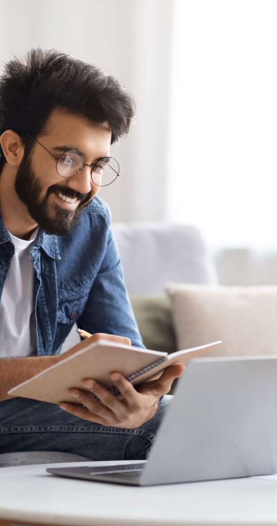 Distance Learning. Young Indian Man Study With Laptop At Home And Taking Notes, Millennial Eastern Guy In Eyeglasses Watching Online Webinar, Sitting On Couch With Computer, Enjoying Remote Education